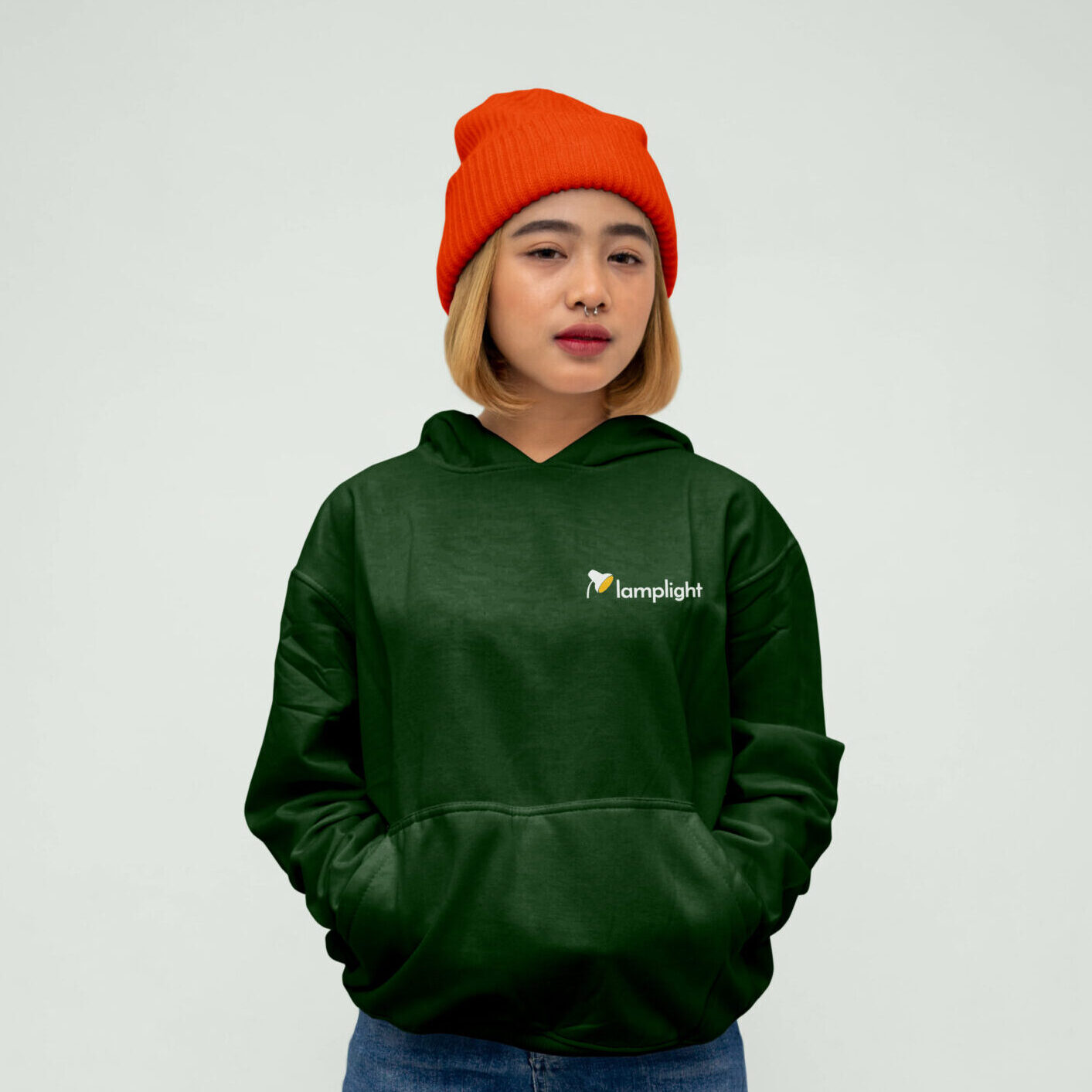 Lady in orange hat wearing Lamplight dark green hoodie mockup featuring new brand identity on front